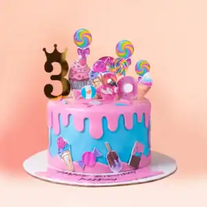 Candy land theme birthday cake in Chennai with pink drip over blue fondant, lollipop, ice cream, cupcake and donut toppers and a gold crown number topper from Cake Square.