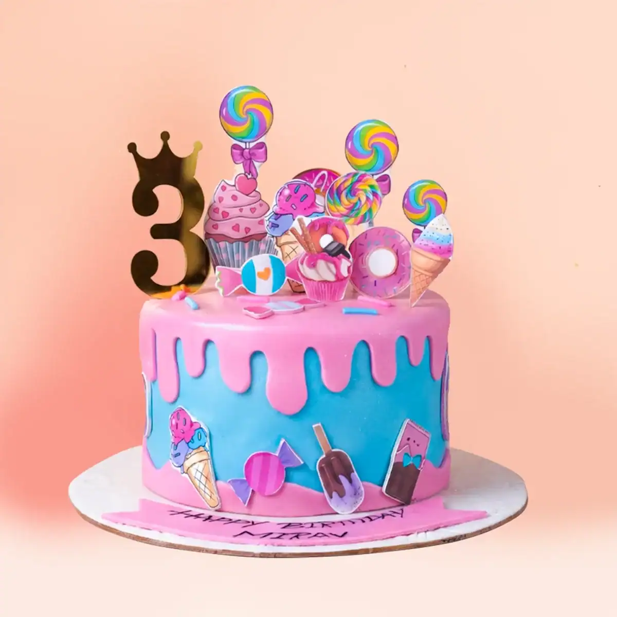 Candy land theme birthday cake in Chennai with pink drip over blue fondant, lollipop, ice cream, cupcake and donut toppers and a gold crown number topper from Cake Square.