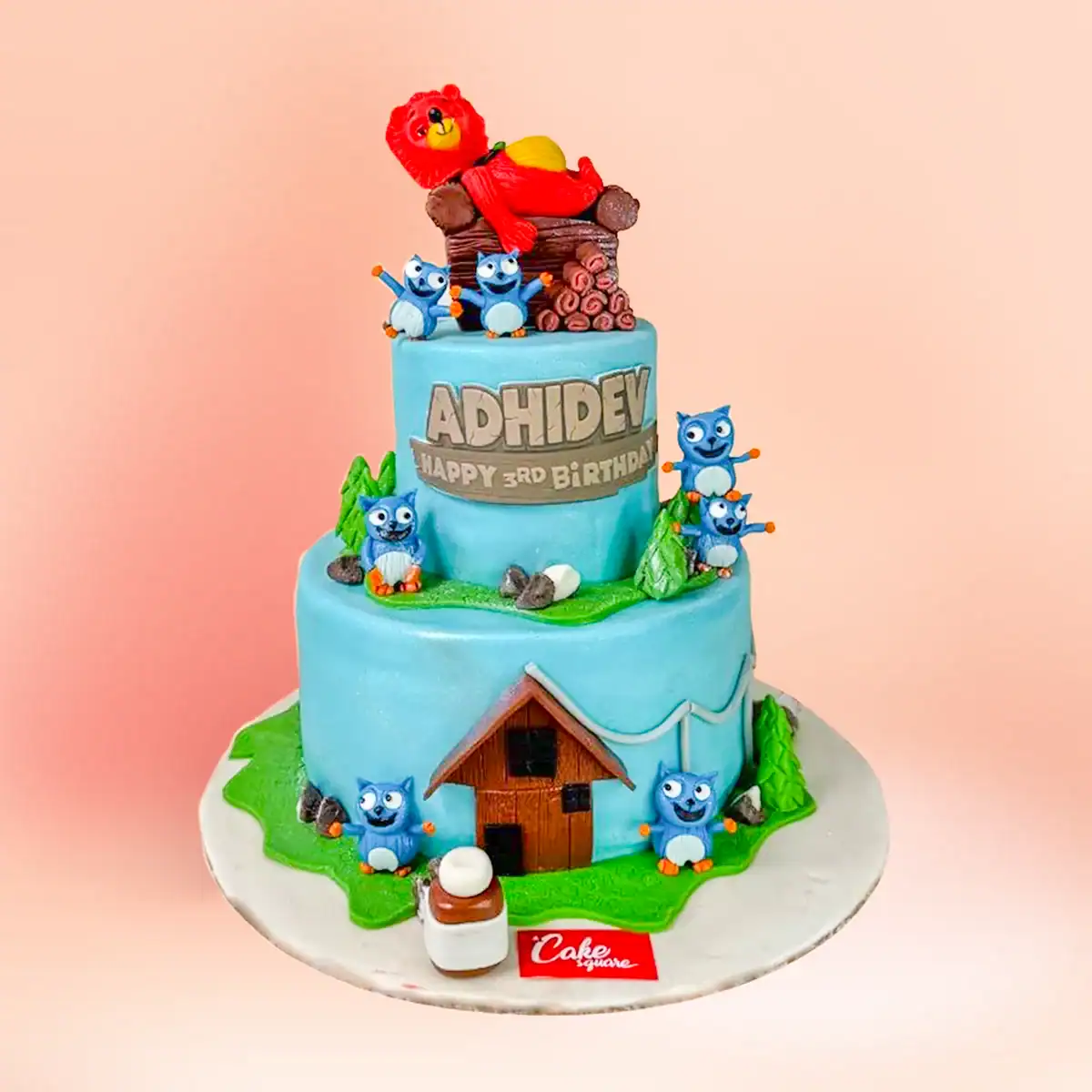 Two tier Grizzy and Lemmings cartoon theme first birthday cake in Chennai with a red bear figure, blue forest friends and a custom name and age topper from Cake Square.