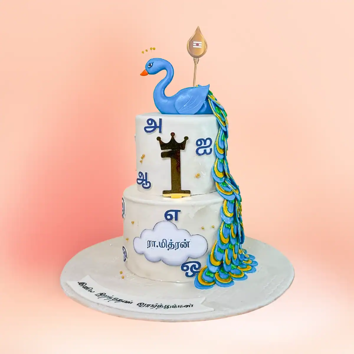 Two tier Murugan theme first birthday cake in Chennai with a blue peacock topper, gold vel and Tamil lettering, available fully eggless from Cake Square.