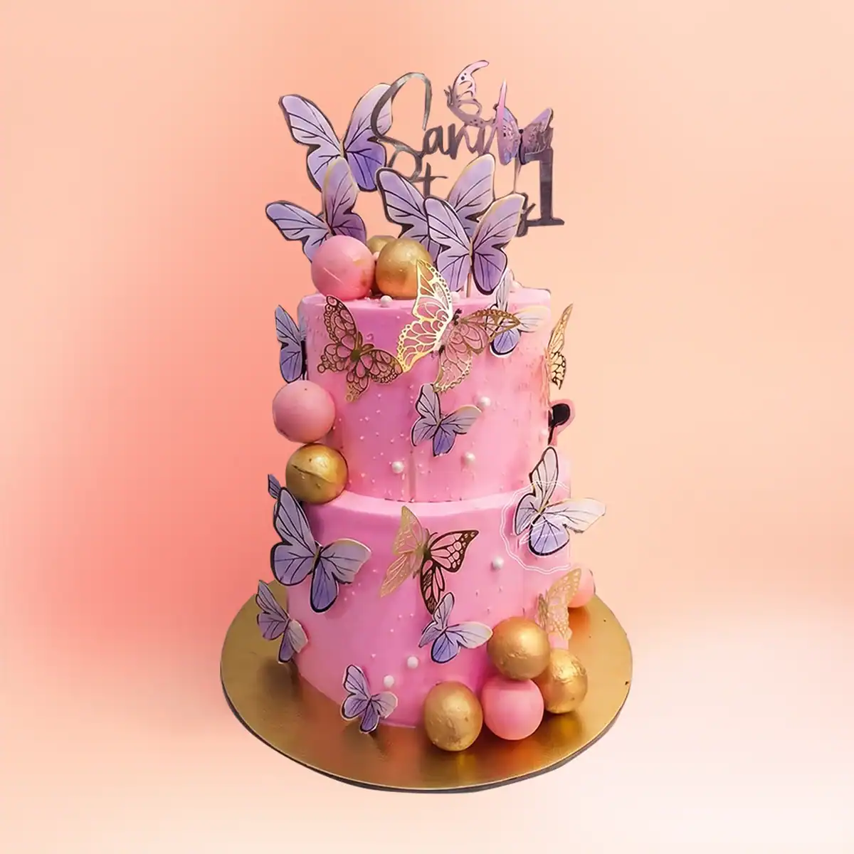 Two tier pink butterfly theme first birthday cake for girls in Chennai with purple and gold butterflies, sugar pearls and a custom name and number one topper from Cake Square.