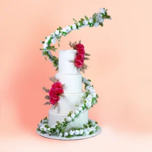Four tier white designer wedding cake in Chennai with a sweeping arch of white roses and greenery over the top, red roses with gold foliage on the tiers and a rose garland at the base from Cake Square.